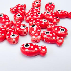 Puede incluir: Una colección de cuentas de plástico rojas y blancas con forma de pez, con lunares blancos y ojos negros. Las cuentas son perfectas para manualidades.