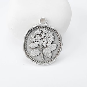 Peut inclure: Pendentif rond argenté avec un motif détaillé d'hortensia. Le pendentif a une bordure texturée avec de petits points et une boucle en haut. La fleur présente des détails complexes, idéal pour la création de bijoux.