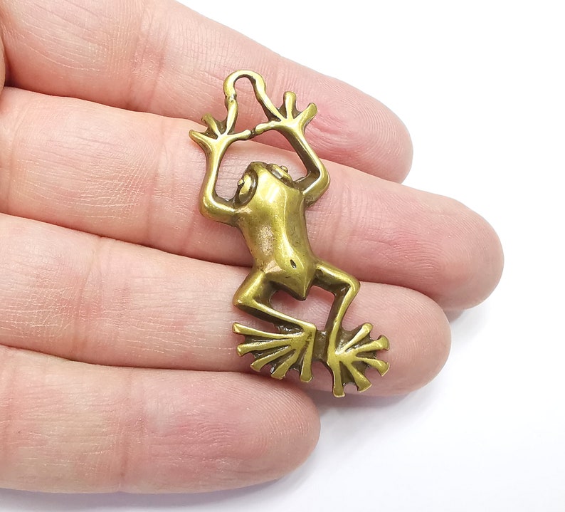 May include: A gold-toned metal frog charm with a loop for hanging. The frog is in a jumping pose with its legs outstretched.