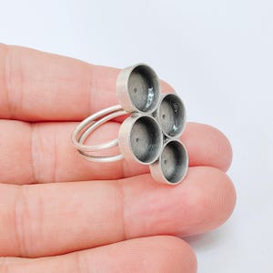 May include: A silver ring featuring five circular settings arranged in a cluster. The ring has a double band design, and the settings are open for adding embellishments. The ring is displayed against a plain background.