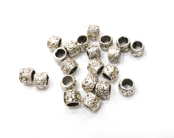 May include: Silver-coloured cylindrical beads with an intricate, swirling pattern. The beads are scattered on a white surface, highlighting their detailed design and texture. These beads are suitable for jewellery making.