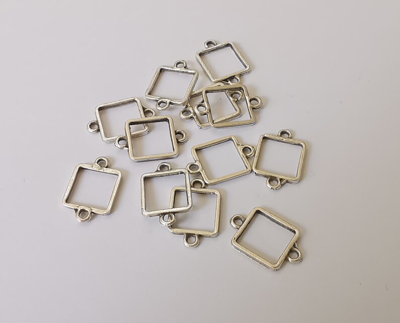 May include: A collection of silver-coloured square connector charms with small loops on each side. The charms have a slightly distressed finish, and are scattered across a light grey surface. These charms are ideal for jewellery making and crafting.