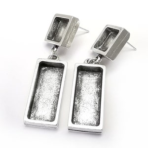May include: A pair of silver earrings with a rectangular design. The earrings have a small square at the top and a larger rectangle hanging below. The earrings are made of metal and have a textured finish.