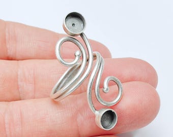 Könnte beinhalten: Ein silberfarbener, verstellbarer Ring mit einem verzierten, wirbelnden Design. Der Ring hat zwei kleine, kreisförmige Fassungen, wahrscheinlich für Edelsteine. Das Design beinhaltet elegante Kurven und ein poliertes Finish.