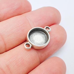 Pode incluir: Uma pequena taça de moldura prateada com duas argolas para fazer joias. A taça tem uma área circular rebaixada para fixar uma pedra ou outro elemento. A moldura é segurada em uma mão.