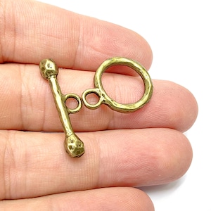 Puede incluir: Un cierre de botón de metal dorado con acabado martillado. El cierre tiene un anillo redondo y una barra con dos anillos pequeños.