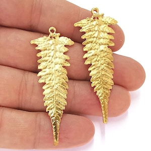 Peut inclure: Deux breloques en forme de feuille de fougère dorées à la surface texturée. Chaque breloque a une boucle en haut pour l'attacher à des bijoux ou d'autres objets artisanaux.