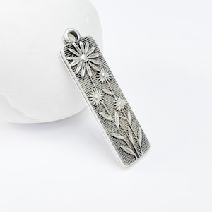 May include: Silver charm with a rectangular design featuring an embossed floral pattern of five white daisies with stems and leaves.