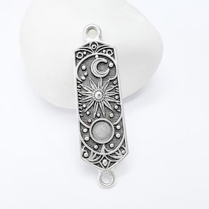 Op de afbeelding: Zilverkleurige rechthoekige hanger met hemels ontwerp. Voorzien van een zon, maan en decoratieve patronen. Inclusief twee lussen voor bevestiging. Ideaal voor het maken van sieraden.