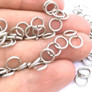 May include: A large quantity of silver jump rings, a type of jewelry finding, are scattered on a hand. The jump rings are small, circular, and have a smooth, polished finish.