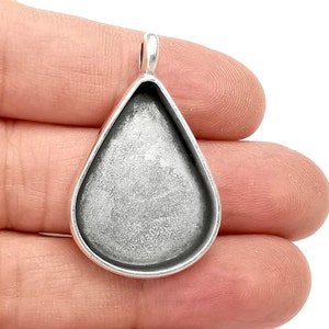 Könnte beinhalten: Silberner Tropfenanhänger mit einer runden, offenen Fassung zum Einsetzen eines Cabochons. Der Anhänger hat oben eine Öse zum Anbringen einer Kette oder Schnur.