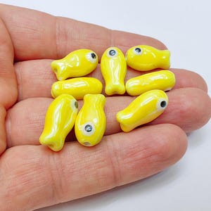 May include: A collection of eight yellow ceramic fish charms with black eyes. The charms are glossy and have a smooth finish.