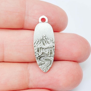 May include: Silver-coloured metal pendant featuring a mountain and river landscape design. The oval-shaped pendant has a small loop at the top for attaching to a chain. The design includes a detailed mountain range, trees, and a winding river.