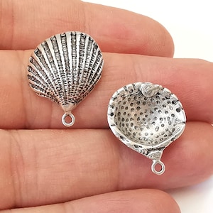 May include: Two silver-toned seashell charms. One charm is a detailed scallop shell, the other is a textured, open-faced shell with a small loop for attaching to jewelry. Both charms have a small loop at the bottom.