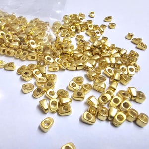 May include: A collection of small, gold-colored metal beads scattered on a white surface. The beads have a rectangular shape with rounded edges and a hole in the center. They appear to be new and unused, possibly for jewelry making or crafts.
