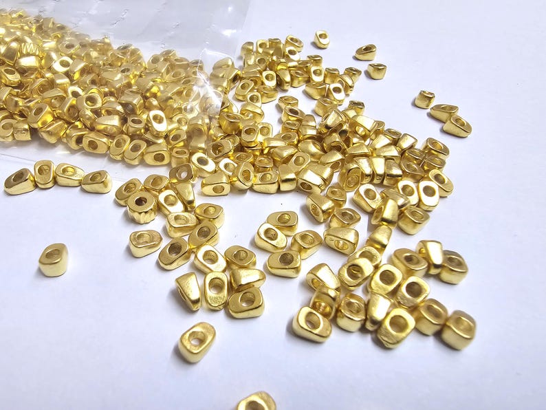 May include: A collection of small, gold-colored metal beads scattered on a white surface. The beads have a rectangular shape with rounded edges and a hole in the center. They appear to be new and unused, possibly for jewelry making or crafts.