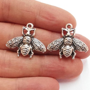 Peut inclure: Deux breloques abeilles en argent avec des ailes détaillées et un corps texturé. Les breloques ont une boucle en haut pour être attachées à des bijoux ou d'autres objets artisanaux.