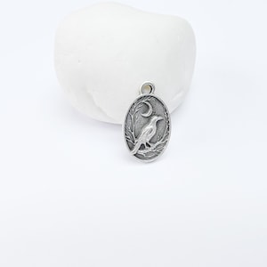 May include: Silver charm featuring a detailed design of a crow perched on a branch with a crescent moon in the background.