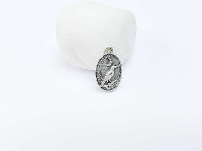 May include: Silver charm featuring a detailed design of a crow perched on a branch with a crescent moon in the background.