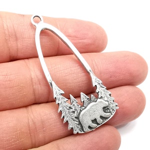 May include: Silver pendant featuring a bear silhouette in a forest scene. The pendant is shaped like an arch and has a loop at the top for hanging.