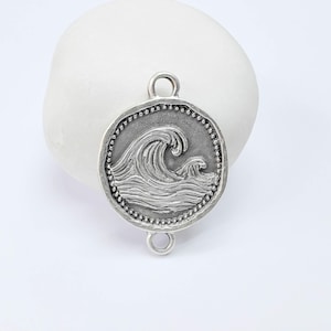 May include: A silver-toned metal charm with a detailed wave design. The round charm features a large, textured wave and a smaller wave, surrounded by a dotted border. It has loops at the top and bottom for attachment.