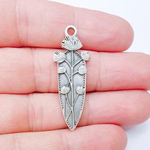 Può includere: Un ciondolo argentato con un design floreale. Il ciondolo presenta uno stelo centrale con fiori e foglie stilizzate, incorniciato da un bordo a puntini. Ha un anello nella parte superiore per il fissaggio a una catena o un cordino.