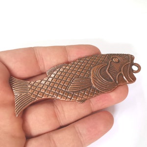 Może przedstawiać: Zawieszka w kształcie ryby w kolorze miedzianym z detalicznym wzorem. Ryba ma fakturowane ciało i wypukłą płetwę. Zawieszka ma pętelkę na górze do przymocowania do naszyjnika lub bransoletki.