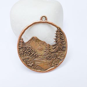 May include: A copper-colored pendant with a mountain and forest scene. The round pendant features a detailed relief of a mountain, trees, and flowers. It has a small loop at the top for hanging.