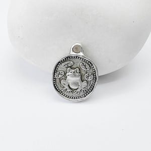 Puede incluir: Un dije redondo plateado con un diseño detallado de rana rodeada de nenúfares. El dije tiene un pequeño lazo en la parte superior para sujetarlo a un collar o pulsera. La rana está centrada dentro de un borde punteado.