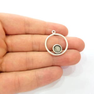 May include: A silver pendant featuring a circular frame with a smaller, circular setting inside. The setting is filled with a textured, grey material. The pendant has a small loop at the top for attaching to a chain or cord.