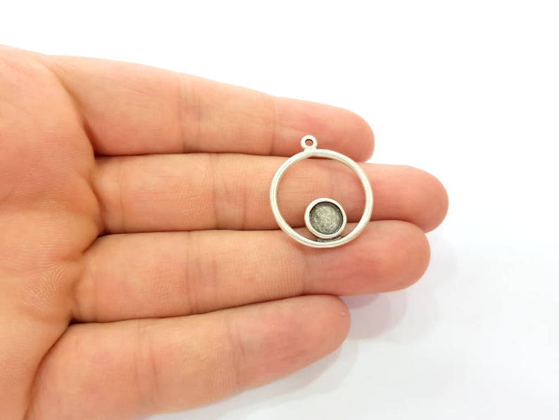May include: A silver pendant featuring a circular frame with a smaller, circular setting inside. The setting is filled with a textured, grey material. The pendant has a small loop at the top for attaching to a chain or cord.