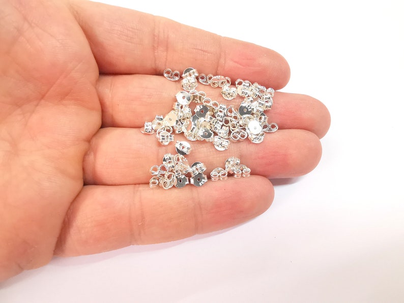 May include: A pile of silver earring backs, also known as earring clutches, in a hand. The earring backs are small and round with a loop on the back.
