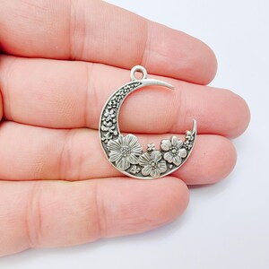 May include: A silver-colored crescent moon pendant with floral detailing. The pendant features a crescent shape with a loop at the top for hanging. The moon is decorated with various flower designs.