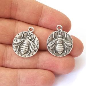 May include: Two silver-toned bee charms with a detailed bee design. The charms are round and have a loop at the top for attaching to jewelry or other crafts.