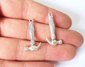 May include: Two small silver-colored hammer charms. The hammer heads are detailed with a curved claw and the handles have a wood grain texture. Each charm has a small loop at the top for attaching to jewelry or other crafts.