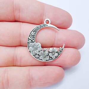 May include: A silver-colored crescent moon pendant with floral detailing. The pendant features three prominent flowers and smaller floral accents along the moon's curve. It has a loop at the top for attaching to a chain or cord. The pendant is held in a hand.