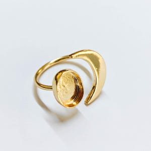 May include: A gold ring featuring an oval setting and a crescent design. The ring has a polished finish and is set against a white backdrop. This piece of jewellery is a ring.