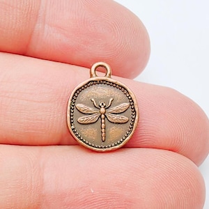 May include: A bronze-colored metal charm with a dragonfly design. The round charm features a detailed dragonfly in the center, surrounded by a beaded border. It has a small loop at the top for attaching to jewelry or other crafts.