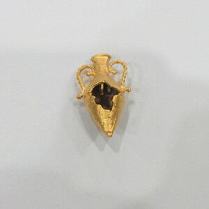 May include: A gold-colored metal pendant in the shape of a vase with two handles. The vase is open at the top and has a textured surface.