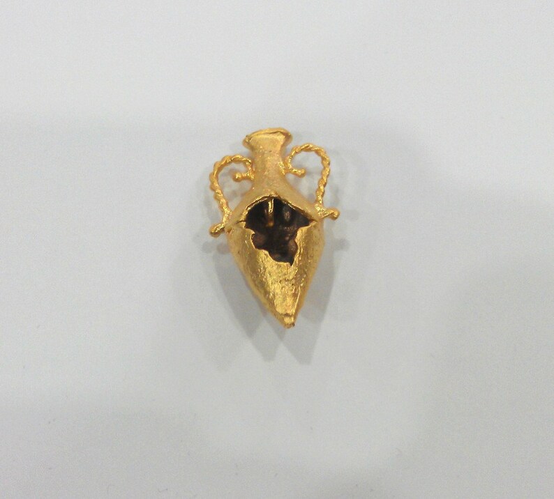 May include: A gold-colored metal pendant in the shape of a vase with two handles. The vase is open at the top and has a textured surface.