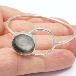 May include: A silver bangle bracelet with a round, open-backed bezel setting. The bezel is designed to hold a cabochon stone or other embellishment.