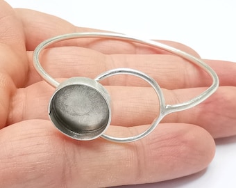 May include: A silver bangle bracelet with a round, open-backed bezel setting. The bezel is designed to hold a cabochon stone or other embellishment.
