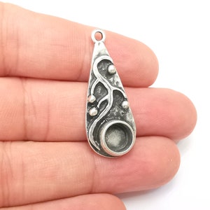 May include: Silver pendant with a teardrop shape and a textured design featuring a branch with small circles. The pendant has a round bezel for a cabochon stone.