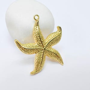 May include: A gold-colored starfish pendant with a textured surface and a loop at the top for hanging. The five-pointed star is set against a white background.