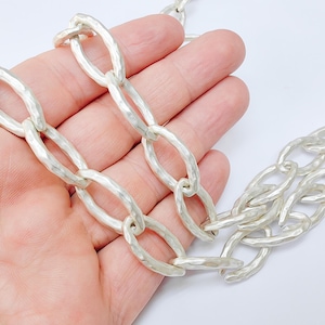 Pode incluir: Um colar de corrente prateado com elos ovais grandes e texturizados. O colar é exibido em um fundo branco, destacando sua aparência de metal martelado. A corrente é uma peça ousada.