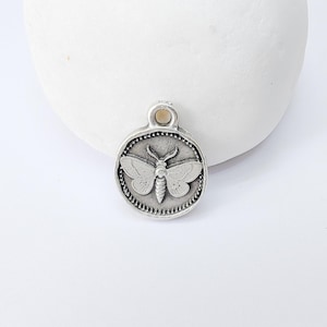 Op de afbeelding: Een kleine, ronde, zilverkleurige hanger met een gedetailleerd motontwerp. De mot is gecentreerd in een kralenrand en de hanger heeft een kleine lus aan de bovenkant om aan een ketting te bevestigen. De hanger is waarschijnlijk van metaal.