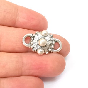 May include: Silver-toned metal clasp with a circular design featuring a raised centre and multiple small beads around the edge. The clasp has two loops on either side for attaching to a chain or cord.