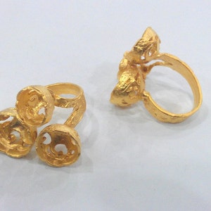 Op de afbeelding: Twee gouden ringen met ingewikkelde ontwerpen. Een ring heeft drie ronde, getextureerde elementen, terwijl de andere een enkel, groter, getextureerd element heeft.