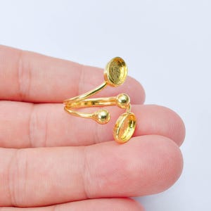 May include: A gold-tone adjustable ring featuring two open circular settings designed for mounting stones or cabochons. The ring has a bypass-style band with two small spherical accents positioned between the two circular bezel cups.
