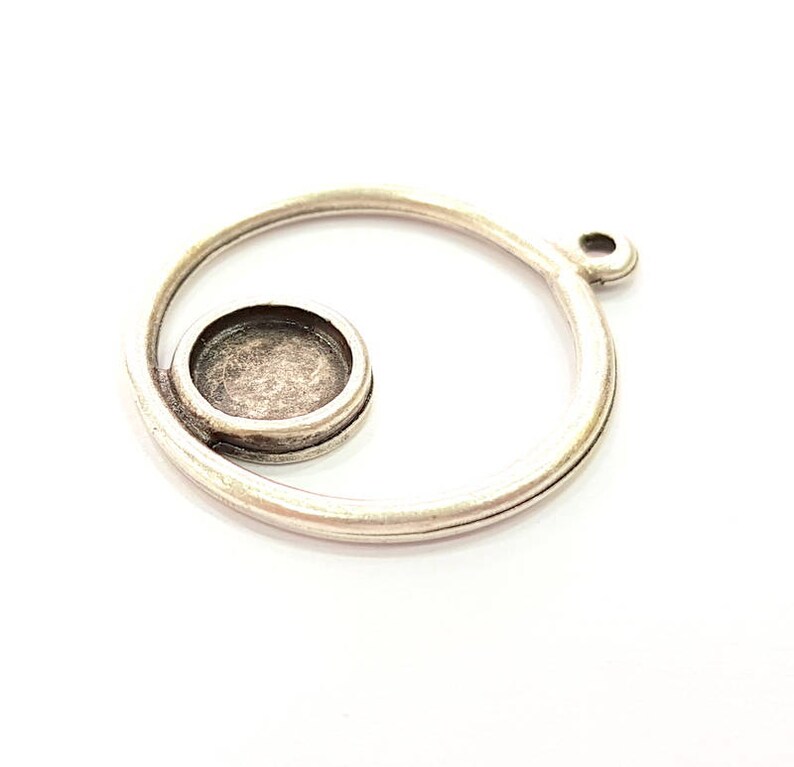 May include: A silver-coloured metal pendant with a spiral design. The pendant has a circular setting for a small stone or embellishment. The design includes a loop for attaching to a chain or cord.